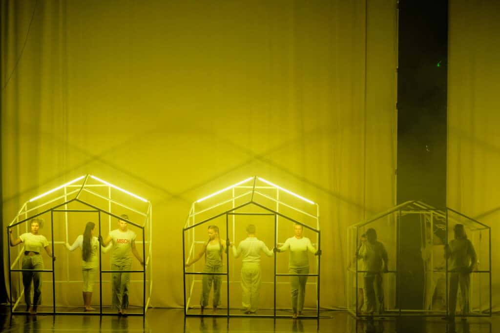 Scena spektaklu. Scena zbiorowa, Trzy grupy osób stoją w trzech rusztowaniach w kształcie domków. Wszyscy ubrani są na biało. Dominuje żółte światło.