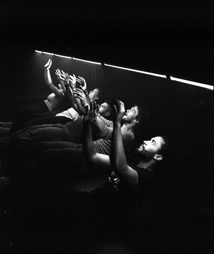 Scena spektaklu. Zdjęcie czarno-białe. Grupa młodych osób lezy koło siebie na podłodze. Wszyscy patrzą w górę. Nad ich głowami co kilkadziesiąt centymetrów widać zapalone świetlówki.