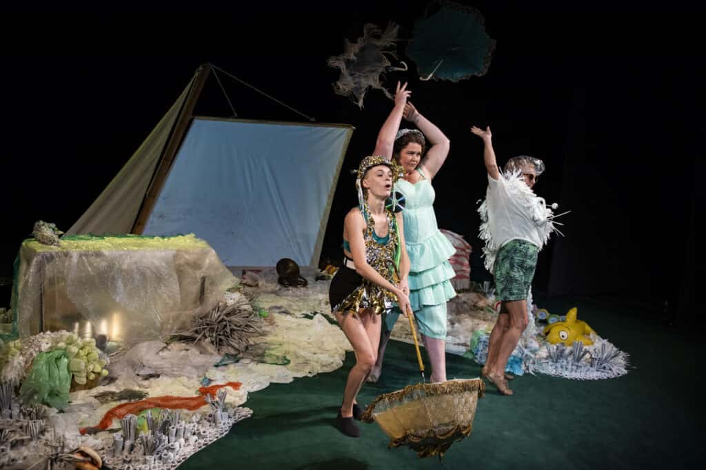 Scena spektaklu. Na scenie stoją trzy kobiety ubrane w letnie kolorowe stroje, kapelusze, okulary przeciwsłoneczne. Kobiety wyrzucają za siebie trzymane parasolki. W tle scenografia przypominająca piaszczystą plażę i nadmorski klimat.