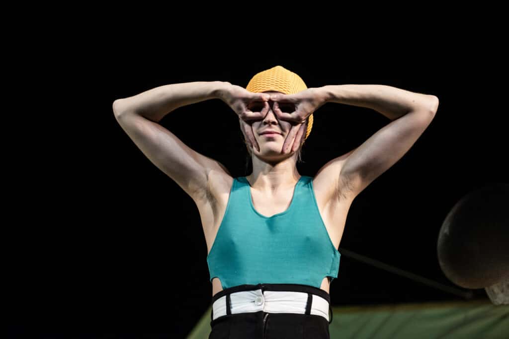 Scena ze spektaklu. WIdzimy od pasa w górę młodą kobietę w stroju kąpielowym i żółtym czepku pływackim. Twarz zakrywa sobie dłońmi, układając palce w kształt okularów lub maski.