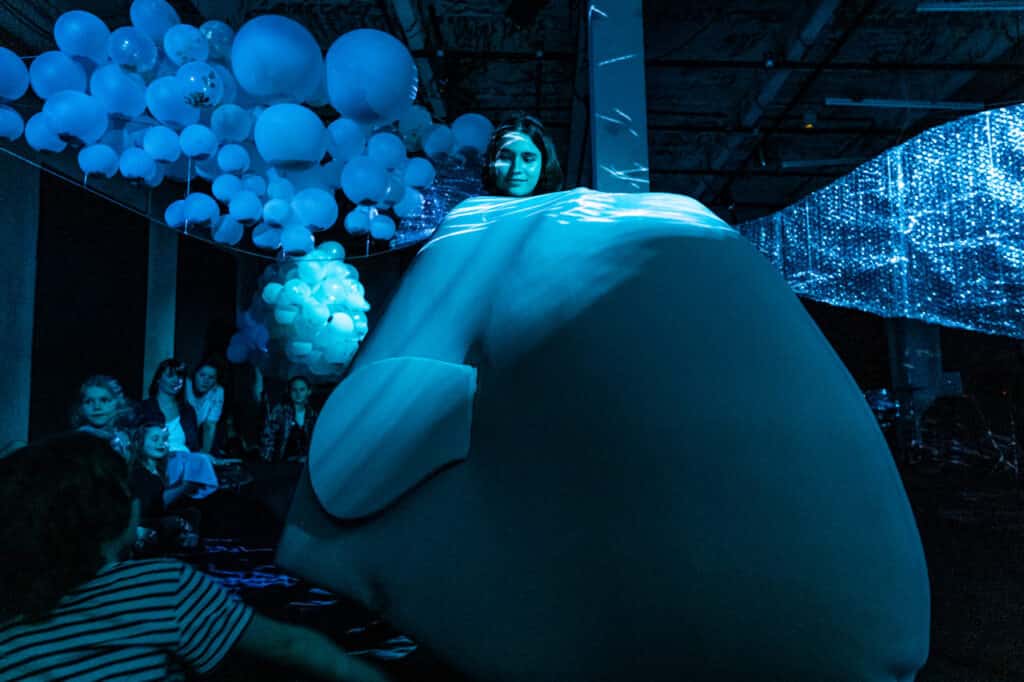 Scena spektaklu. Młoda dziewczyn siedzi na samej górze wielkiech poduszki. Poduszka ma kształt wieloryba. Cała scena oświetlona jest na niebiesko.