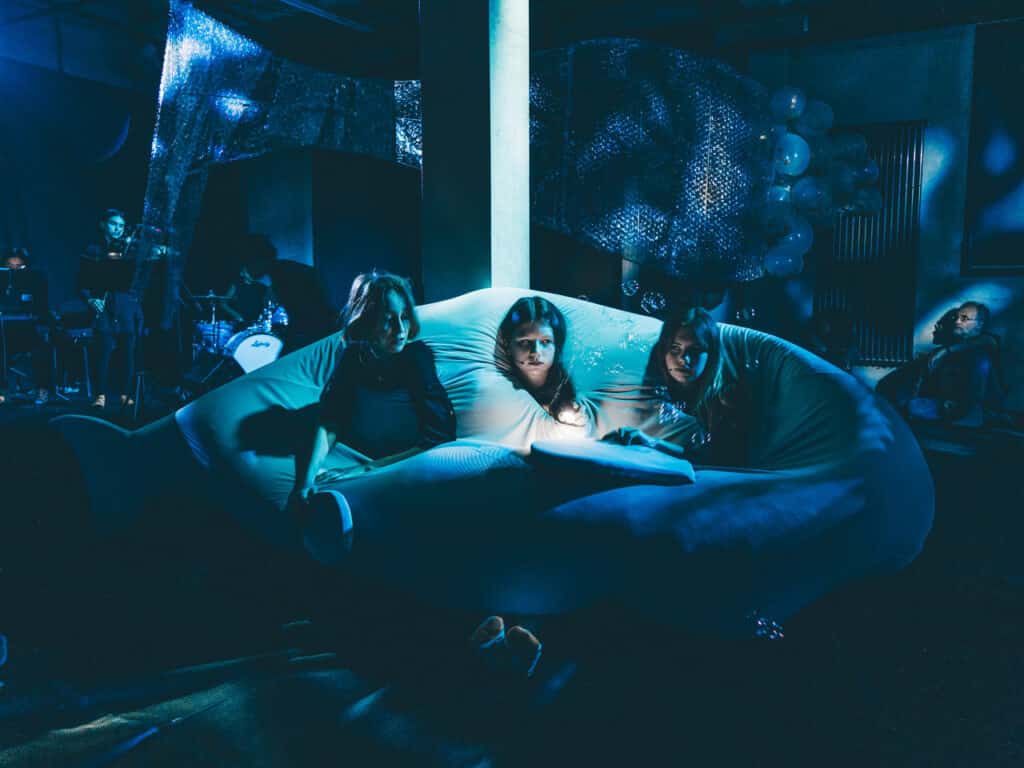 Scena ze spektaklu. Na wielkiej miękkiej poduszce siedzą trzy dziewczyny. Jedna trzyma w ręku małą latarkę. Za nimi, w kącie sali widać zespół muzyczny. Całość sceny spowita w zielono-niebieskim świetle.