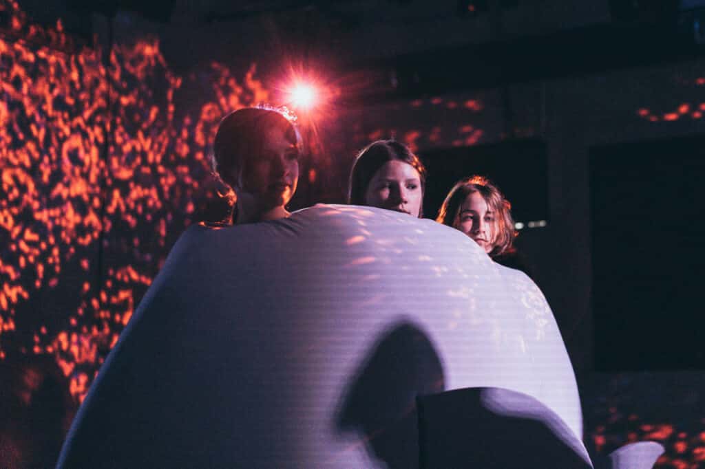Scena z próby do spektaklu. Na wielkiej poduszce w kształcie wieloryba siedzą trzy dziewczyny. Sylwetki postaci oświetlone są na fioletowo, twarze oświetlone są w czerwone plamki.