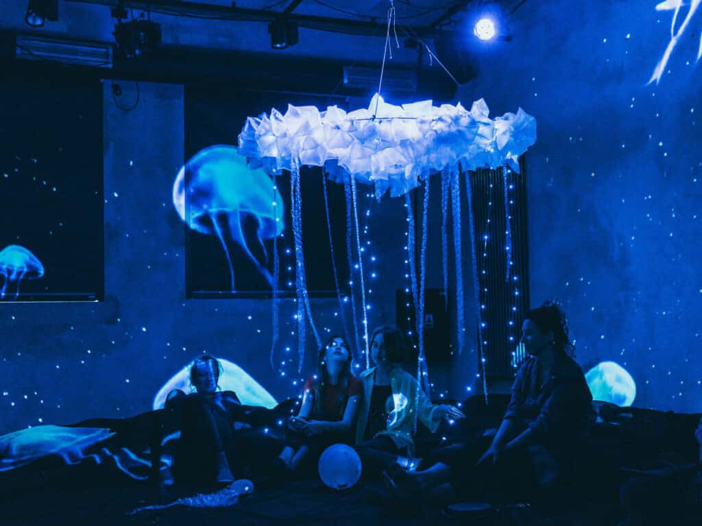 Scena z próby do spektaklu. Na podłodze siedzą młode dziewczyny, nad nimi wisi żyrandol z białej krepiny, z którego zwisają sznurki białych młych światełek choinkowych, całość wygląda jak meduza. Na ścianach widać projekcje z pływającymi meduzami.