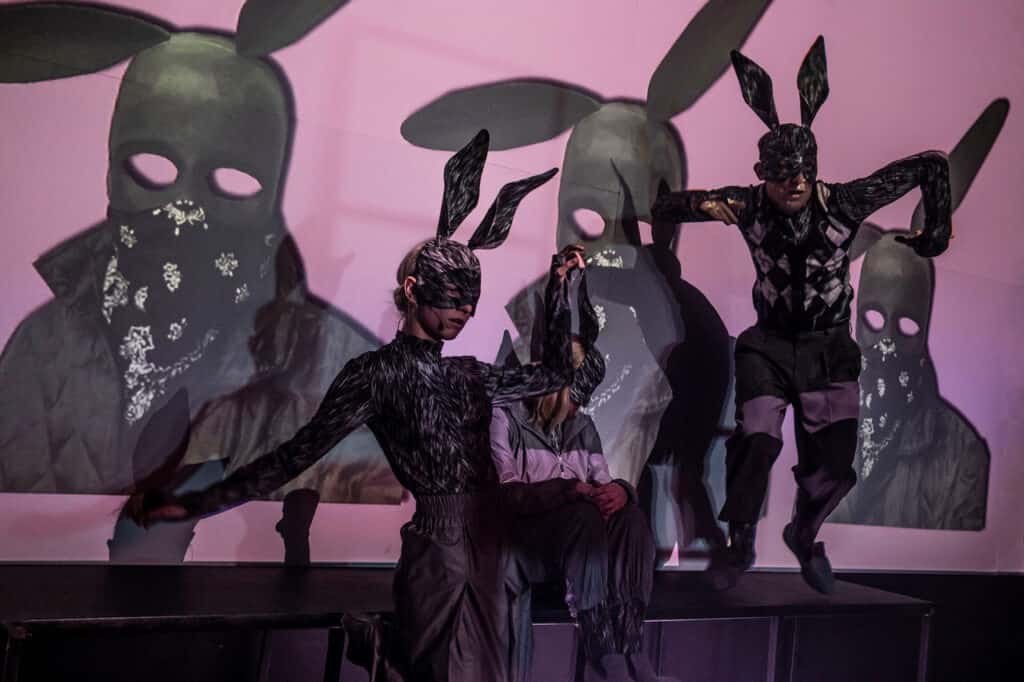 Scena spektaklu. Dwie aktorki w maskách królików, jedna stoi, druga siedzi i patrzy na ekran na którym wyswietlona jest maska królika na różowym tle. Obok trzevi kator także w masce królika skacze z podestu.