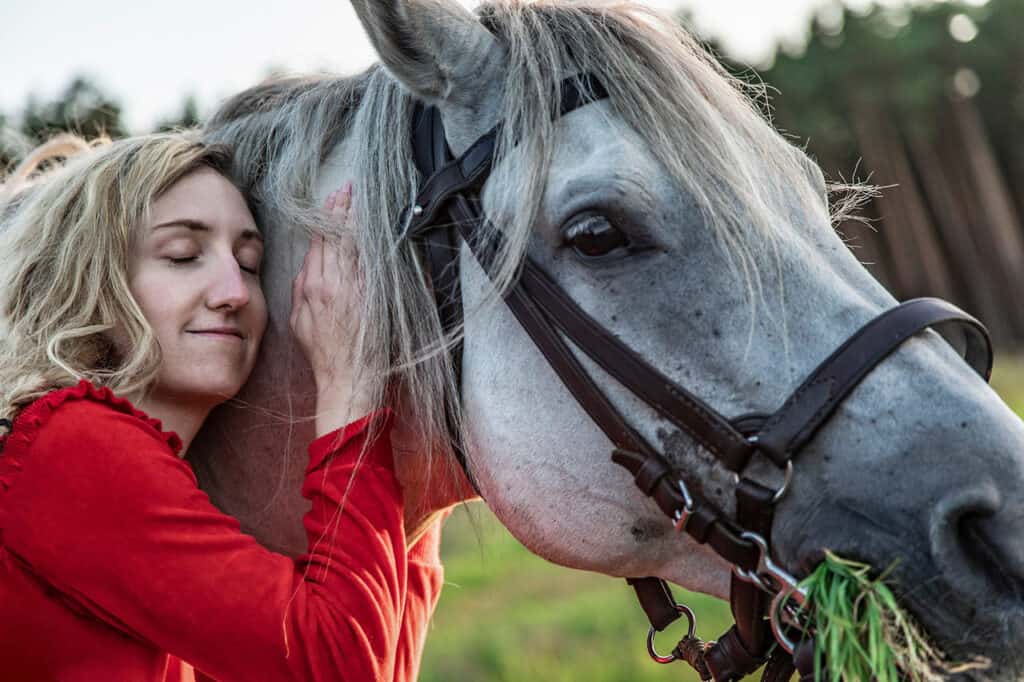Scena w plenerze. Młoda kobieta w czerwonej sukience stoi przy śniadym koniu, przysuwa twarz do jego grzywy, obejmuje go, ma zamknięte oczy i uśmiecha się. Koń ma na sobie uzdę, w pysku trzyma trawę. W tle widać wysokie drrzewa.